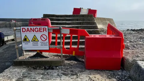 Dorset Council Red barriers and signs in front of stone steps leading to the high wall of The Cobb, with the sea in the background