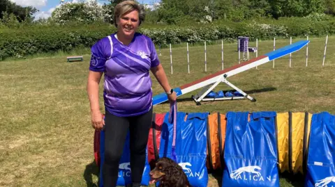 A woman is standing in a field with her dog in front of a blue seesaw. The woman has short brunette hair and is wearing a purple top.