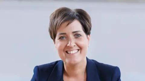 A head shot of a woman with short brown hair in a navy blazer smiling to the camera. The background is white.