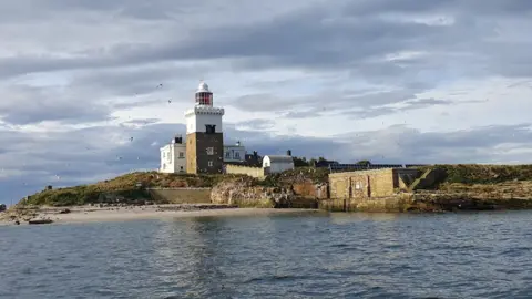 RSPB Coquet Island, taken from the sea. Navy blue waves surround the small island off the coast of Northumberland. There is a lighthouse and a number of small, brick buildings.