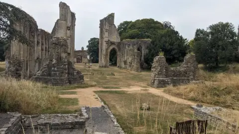 Glastonbury Abbey The photo shows the ruins of Glastonbury Abbey. A grassy lawn fills the middle of the image, with stone arches to its left and right. The grass is largely brown.