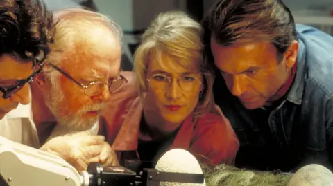 Getty Images From left to right, actors Jeff Goldblum as Dr. Ian Malcolm, Richard Attenborough as John Hammond, Laura Dern as Dr. Ellie Sattler and Sam Neill as Dr. Alan Grant, watching a robotic arm handle the dinosaur eggs in a scene from the film 'Jurassic Park', 1993. 