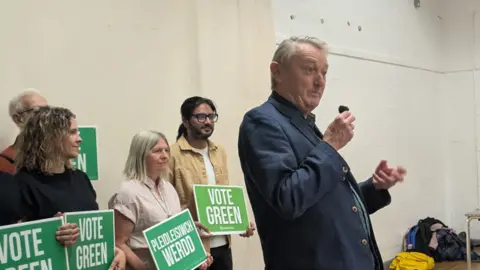 Anthony Slaughter in a blue suit holding a microphone with four people behind him holding vote Green signs in English and Welsh.
