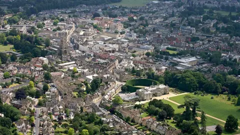 An aerial view of Cirencester, a town in the Cotswolds. It is full of Cotswold stone buildings and a stately home with a large lawn can be seen.
