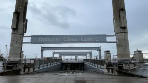 The entrance of the National Glass Centre in Sunderland. A large grey sign is suspended between two high cement pillars. A sloped path leads to the glass doors of the building. The fenced off roof of the building is visible in the distance.
