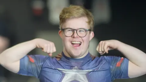 Jamie Niblock/BBC Edward Wagland with blonde hair smiles widely at the camera while tensing his bicep muscles in his arms. He is wearing a blue top with a silver star in the middle of it as well as black glasses.