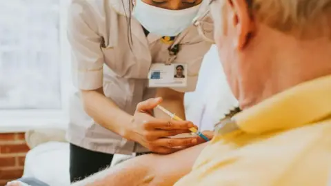 A medical professional in a white coat is injecting a seated man in his arm. He is wearing glasses and a yellow shirt