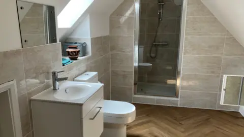 JOANNA TAYLOR/BBC A clean en ensuite bathroom with grey tiled walls, a white sink and toilet, mirror and shower built into the wall. The floor is wood panelled. There is also a sky light above the toilet and a small vase on a windowsill underneath it.