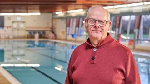 A man wearing glasses and a red top stands next to a swimming pool. 