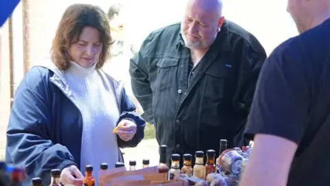 BBC/ITV Cymru Wales Ruth Jones seen visiting a market with Steve Speirs. She is wearing a blue rain jacket and a white turtle neck jumper and is looking at bottles of sauces at a stall.