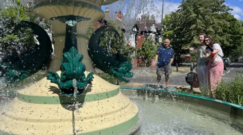 A yellow and green fountain spurting out water. In the back there are people looking into it. There are houses and a large tree in the back.