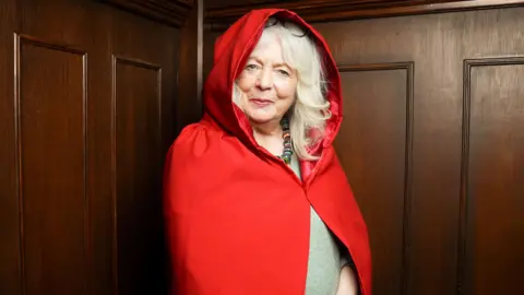Alison Steadman as secret traitor Fiona, she is smiling at the camera conspiratorially and wearing a red hooded cloak.