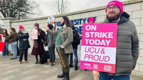 A picket line at University of Aberdeen with a striker holding up a cardboard sign that reads: "On strike today / UCU & proud".