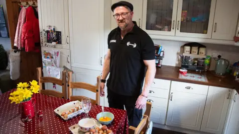 BBC News A person stands in a brightly lit kitchen beside a table, where there are several bowls and egg cartons. The person is wearing a dark polo shirt with a small weightlifting graphic and text on the chest. 