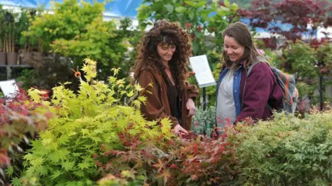 Two women are standing among rows of shrubs and are smiling. The woman on the left, who has long hair and is wearing a brown jacket, is touching some leaves. 