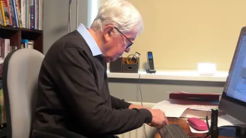 Nigel Thomas is pictured at his computer in his study. Behind him are dark wood bookshelves which are full of books. In front of him is a computer and keyboard, and a pink glasses case. He's wearing a black or dark grey jumper with pale blue shirt collar showing. His hair is white and he has glasses.