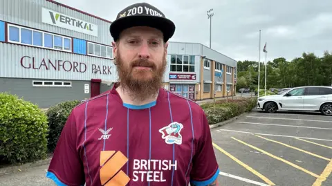 A man stands in the car park of a football ground with the name "Glanford Park" written on the side of a stand. He has a brown beard, piercings and wears a black cap and claret Scunthorpe United football shirt with blue trim and the sponsor name "British Steel" written on the front.