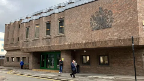 Chelmsford Crown Court, which is a red brick building that has green doors at its entrance. There are three people milling about outside the building.