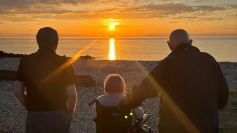 Donald Morrison the backs of two men and a woman, who is in a wheelchair, over looking the water with an orange sun setting in the background.
