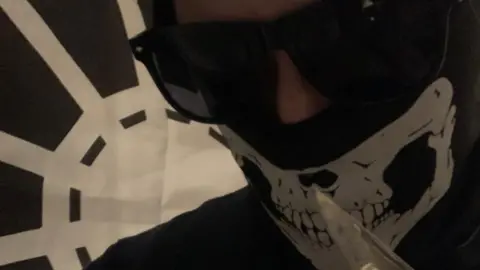 Met Police Alfie Coleman wearing a balaclava with a skull on it and sunglasses while holding a knife. A black and white flag - which looks like a white spider's web - is pinned to a wall behind him.
