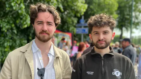 Two students standing side by side outside a university campus. Both with long curly hair and beards.