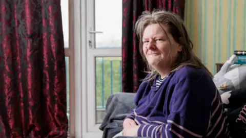 Image shows Ruth, a middle-aged woman with shoulder-length dark hair, sitting on her sofa in her living room, which is now organised and clean. 