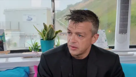Former PC Lorne Castle sits giving a TV interview. He has short dark brown hair with grey flecks. He is wearing black blazer with a black round neck t shirt and he is sat on a blue sofa.