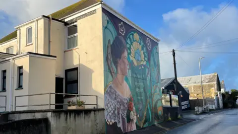 Newquay Business Improvement District The mural is on the side of a building overlooking a main road. It shows Lady Molesworth-St Aubyn in profile wearing a lace top and a flower brooch in the WI’s colours. A central banner reads Newquay WI, Inspiring Women since 1918 framed within a triple-arched Art Nouveau window. There are also pilchards and the Huer’s Hut in the mural.