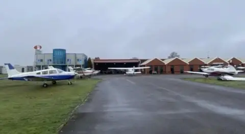A number of planes parked on grass alongside an area of tarmac with a number of brick buildings in the background.