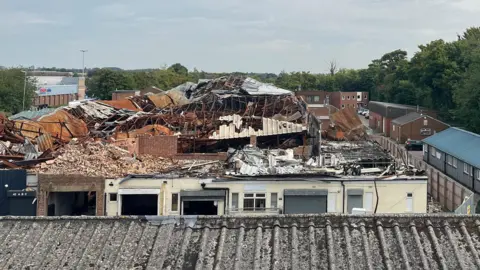 Tony Fisher/BBC An aerial view of an industrial estate which has been destroyed by fire. Crumpled roofing and loose bricks sit on top of a rectangular structure which is surrounded by hoardings. There are intact buildings outside of the hoardings on the edge. A corrugated roof is in the foreground.