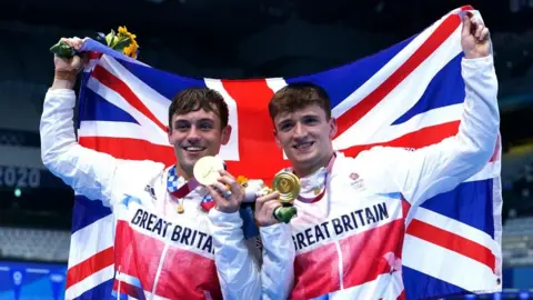 PA Media Two young men with brown hair wearing Great Britain track tops holding up a big union jack behind them while holding a gold medal each in their other hands