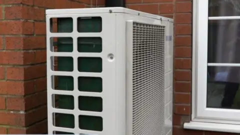 The heat pump is a white machine which is attached to the outer brick wall of the house.