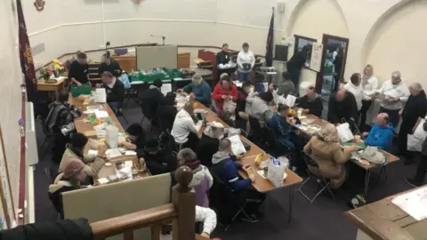 Newbury Soup Kitchen A crowd of people sat around tables eating with people serving them food on the edges of the room