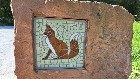 Eden Rivers Trust A fox mosaic is displayed on a sandstone wall. The fox is pieced together by small orange pieces with a green background. The large sandstone plaque stands on a walking trail.