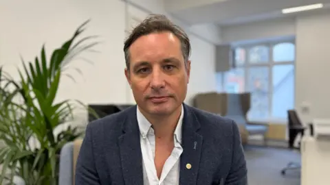Rollo de Sausmarez, Committee Member, Better Journeys Project in the Channel Islands, in an office wearing a blue blazer and white shirt.
