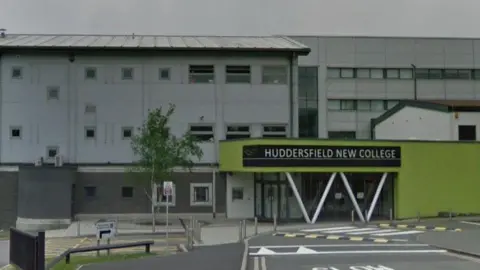 Google A grey four storey building with a lime green single storey entrance. A sign above the glass doors reads 'Huddersfield New College'. A road with speed bumps curves to the right of the entrance.