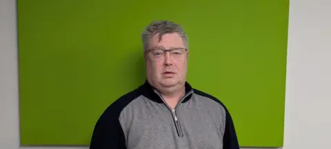 Gavin Harrower Gavin has short grey hair. He is wearing glasses and a grey top. He is standing in front of a green wall.