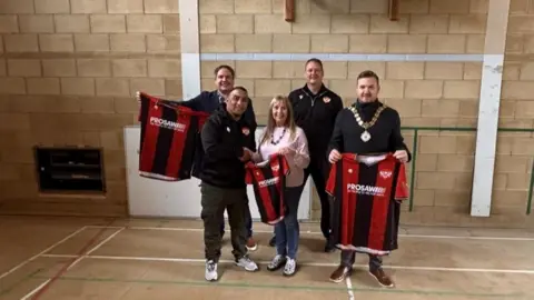 Kettering Town Football Club Four men, all dressed in black tops, and a woman, in a pink jumper, stand in a sports hall holding up three red and black football shirts. 