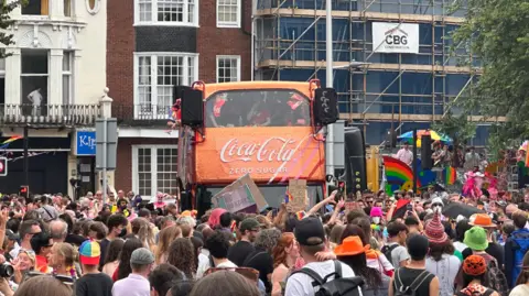 Hundreds of people stood in front of an orange double-decker bus with the Coca-Cola logo on it. Behind are houses and scaffolding