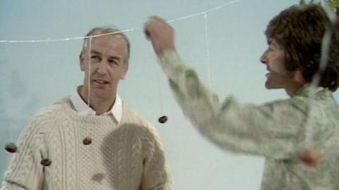 Inside the Blue Peter studio: World Champion of Conkers Peter Midlane on left in an aron sweater.  Blue Peter presenter Peter Purves in a white and green shirt, on the right holding onto a string washing line with conkers on strings hanging off it.