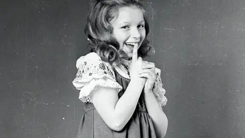 A black and white photo of a young Lena Zavaroni smiling with her finger at her mouth. She is wearing a pinafore and a floral trimmed blouse with puff sleeves.