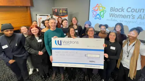 BCWA A group of people all smiling and standing around a large blue and white cheque that says "Waldrons solicitors" in the top left corner. The group include several women and a couple of men. They are indoors and stand in front of a screen showing the Black Country Women's Aid logo and text of their title.