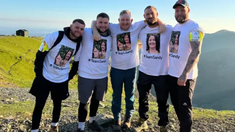 Compton Care Mr Broadhead standing with his arms around friends on Snowdon. They are all wearing white T-shirts with his wife's picture on 