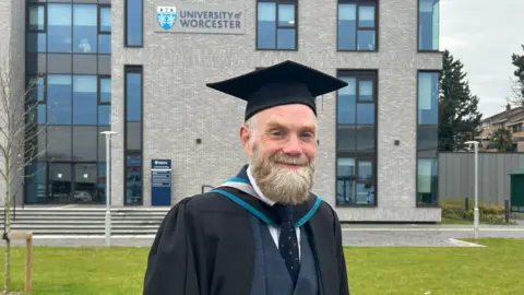 A man with a bushy light/dark grey beard stands smiling at the camera. He is wearing a black mortarboard and a mid-blue suit with a dark blue tie under a black graduation gown. Behind him is a grey brick building with a sign reading: "University of Worcester".