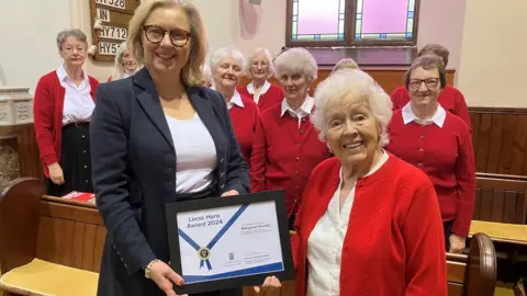 A woman with long blonde hair wearing glasses a white T-shirt with a suit-type jacket presents a certificate to Rita who has white hair and is wearing a white blouse and red cardigan