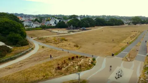 BCP Council An aerial image of Whitecliff in Poole, showing the people walking along grassy paths and people cycling on constructed cycle routes. The grass appears brown and there are large green trees in the distance with houses seen a little bit further than that.