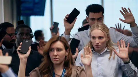 Amazon Content Services LLC Office staff appear with their hands up and phones in hand. They appear with shocked looks on their faces. Sophie Turner's character is wearing a crumpled blue shirt and Archie Madekwe is stood behind her in a similar outfit.