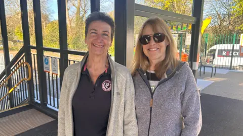 Mags and Penny pictured smiling while stood on a train platform. One of the ladies has brown short hair, wears a blue polo t-shirt and grey jacket on top. While the other has shoulder length blonde hair with a side fringe, she wears large dark circular sun glasses, a grey t-shirt and blue zip jacket.
