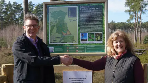 Norfolk Wildlife Trust Eliot Lyne, who has greying light brown hair and is wearing a black coat and blue shirt, shakes hands with Sue Holland, who has light brown shoulder-length hair and is wearing a patterned scarf and black jacket. They are standing in front of a Broadland Country Park sign.