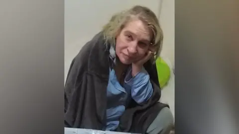 Cambridgeshire Police Julie Buckley: a blonde-haired woman sitting with her head resting on her left hand, She and is wearing a blue shirt and has a grey top hanging over her shoulders.